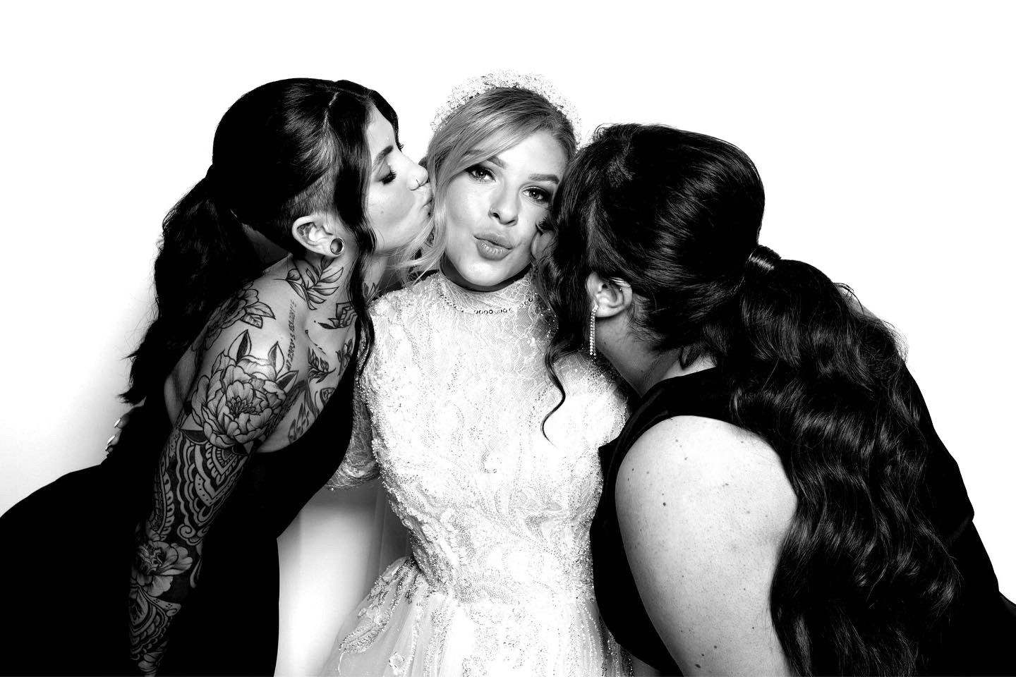 Black-and-white Glam Booth portrait: a bride kissed on both cheeks by two friends